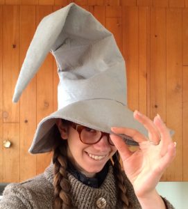 Photo of Paige wearing a large grey wizard's hat. She is wearing glasses and her hair is in two braids. She is smiling and tipping the brim of the wizard hat at the viewer.