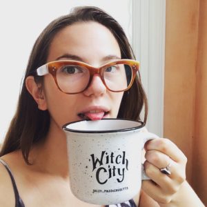 Photo of Paige wearing brown glasses and holding a large white mug that says Witch City in fancy curved letters. Underneath, in smaller letters, it says Salem, Massachusetts. Paige is smiling and sticking her tongue out a little bit.