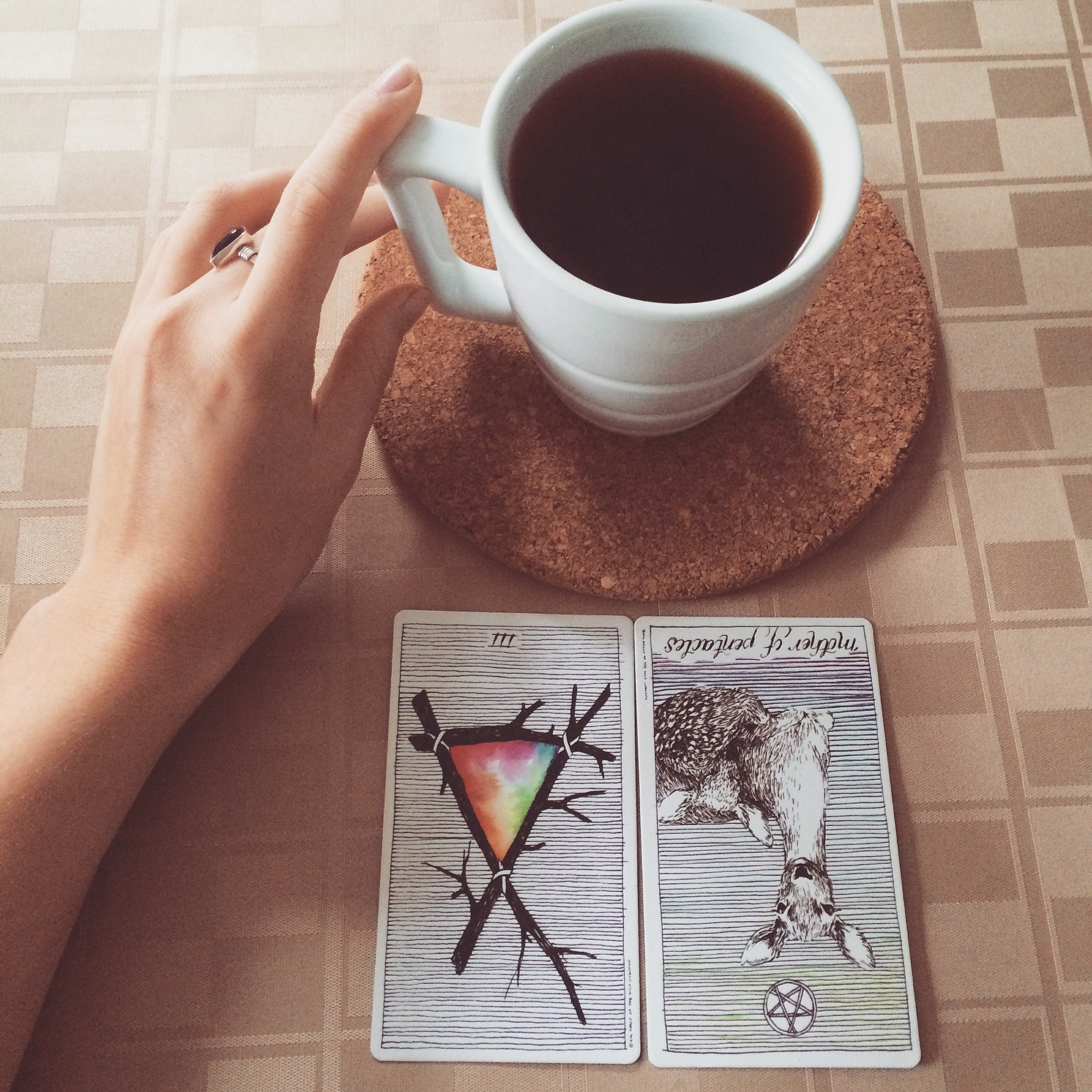 a hand touching a full cup of black tea with the three of wands and the mother of pentacles reversed laid out before the cup