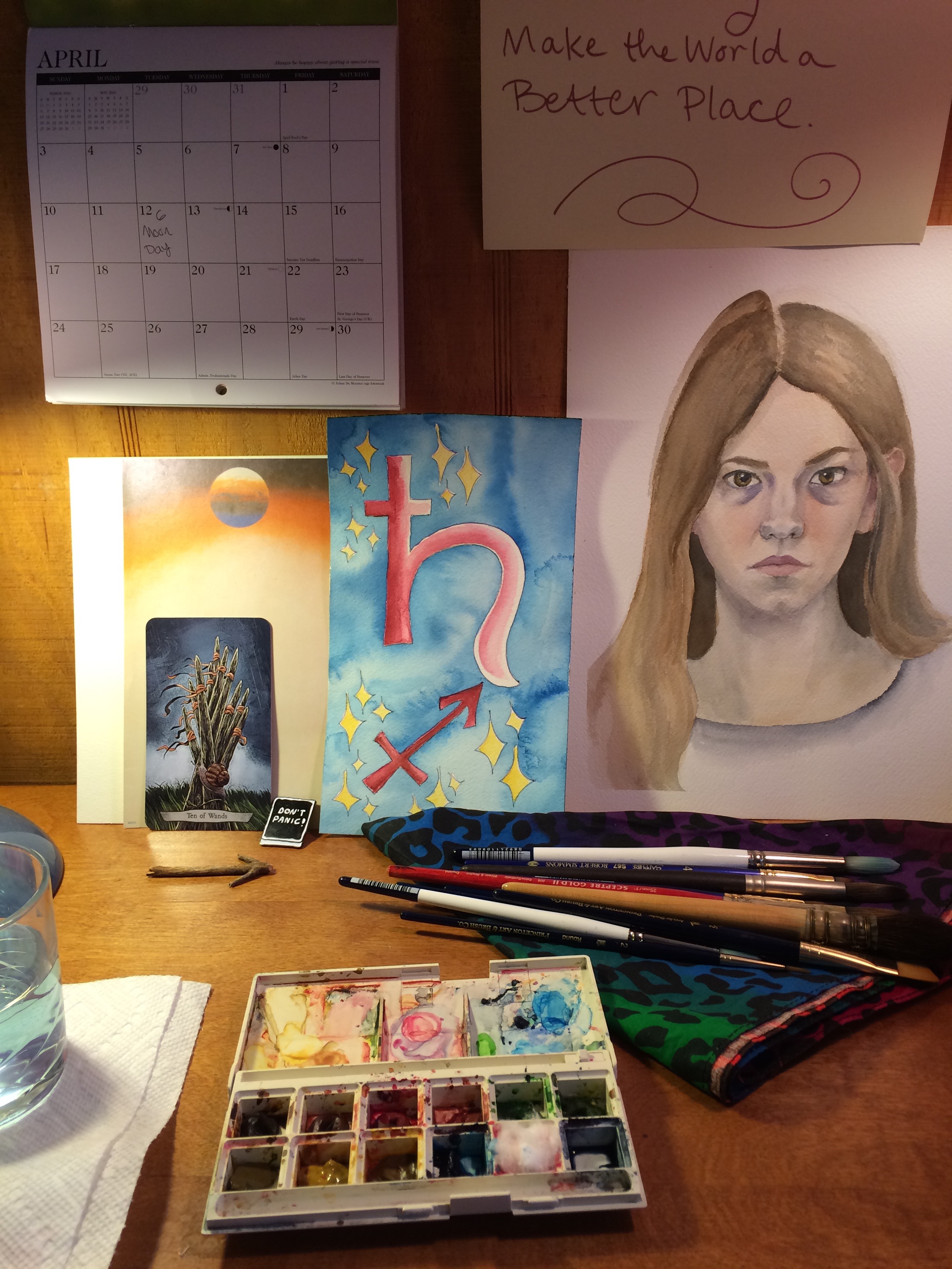 a watercolor palette open on a desk, a group of brushes laying on a rainbow leopard print kerchief beside it. on the wall behind the desk is propped an old unfinished watercolor self-portrait, a watercolor image of the Saturn and Sagittarius sigils on a blue background, a calendar open to April, and the Ten of Wands tarot card leaning against a blank piece of stationary topped with a planet. a glass of painting water is visible on the left.