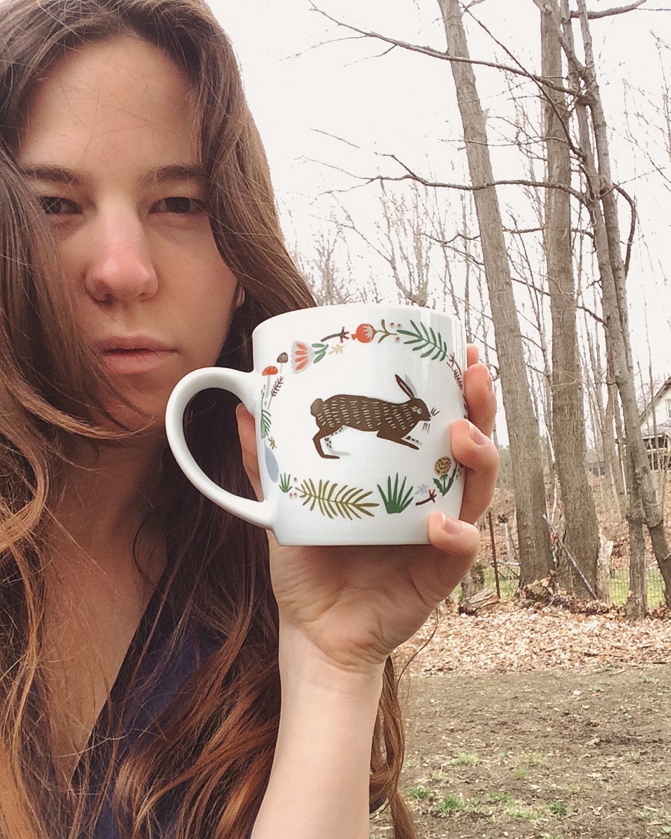 Paige looks at you; bare trees behind steady gaze behind rabbit mug of tea