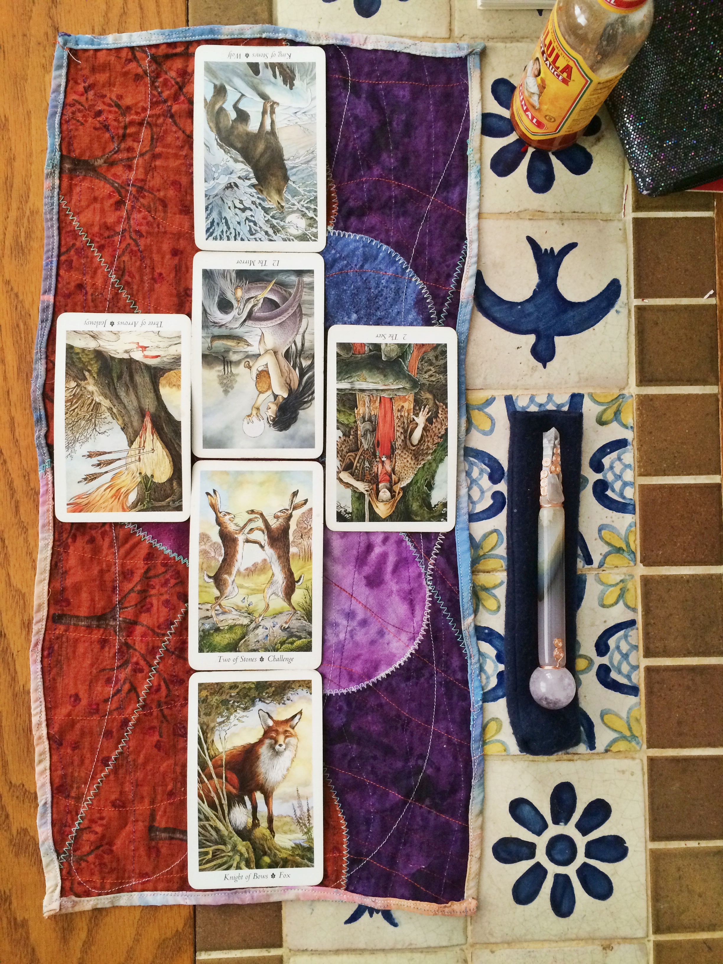 six tarot cards are laid out in a long, skinny cross pattern. to the right is a crystal wand atop a dark blue cloth sheath. beneath it are Mexican tiles of birds and flowers. a bottle of Cholula hot sauce lurks in the background