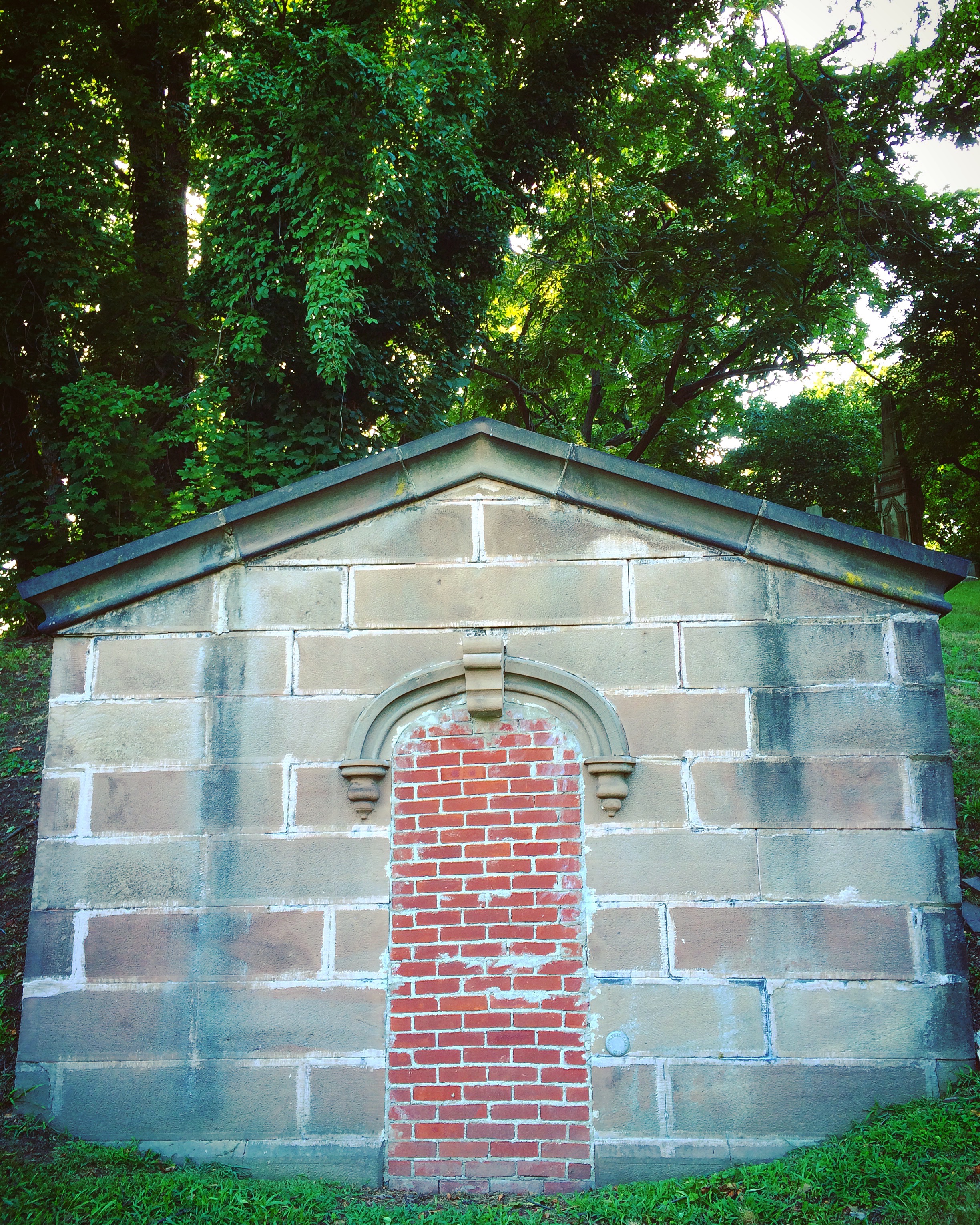 a stone mausoleum with a peaked roof stands before a wall of green trees and overgrowth. its door, beneath a carved arch, is bricked over like some creepy Beetlejuice business