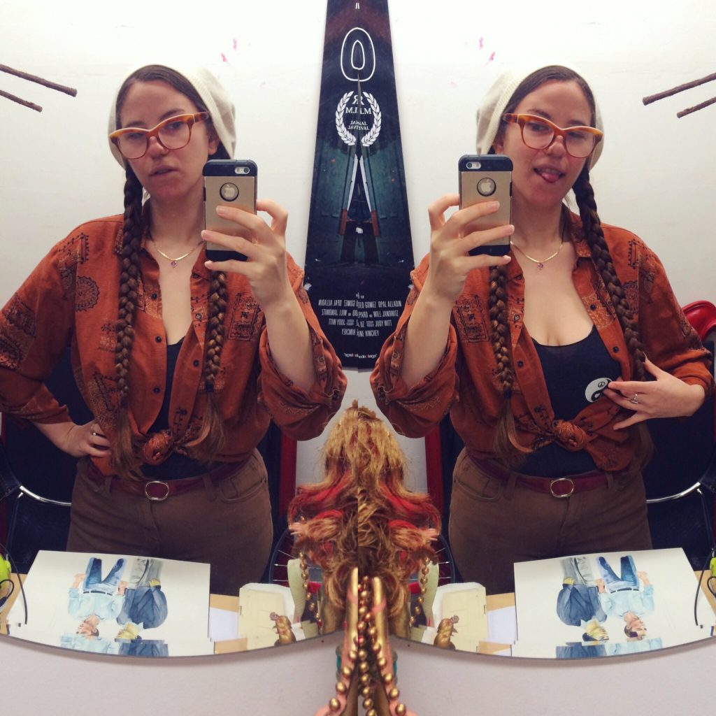 a mirror image of paige wearing a white beanie, glasses, and twin braids taking a selfie in the mirror and pulling back her outer shirt to reveal a secret yin yang on the shirt beneath