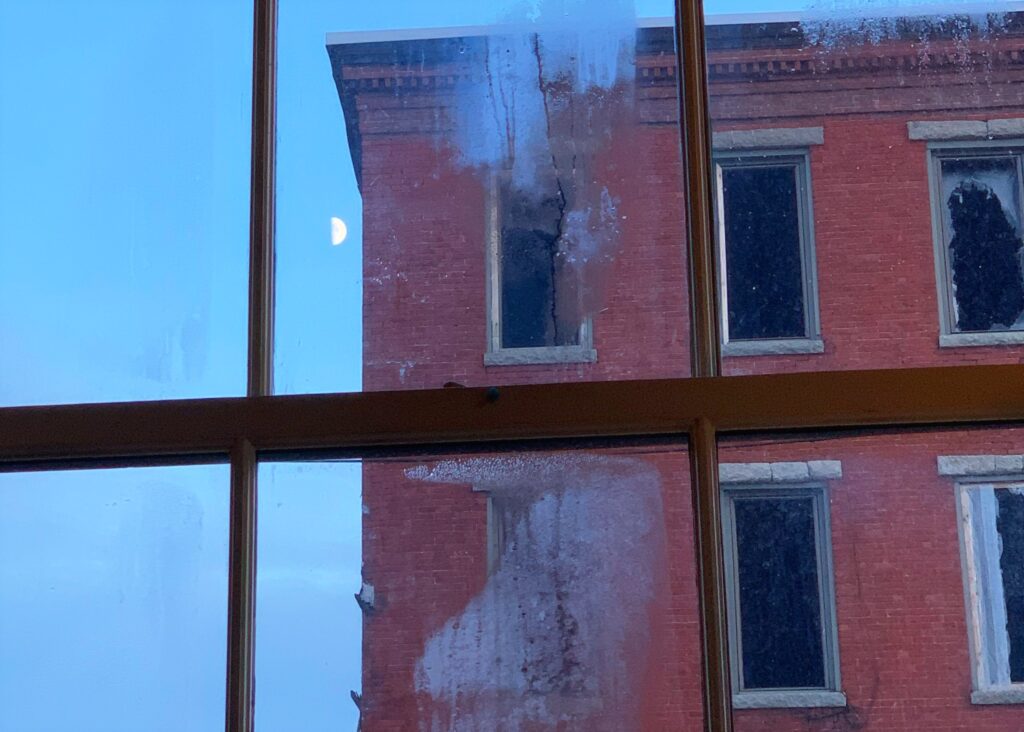 a half moon is visible in the sky next to a brick building with tall thin dark windows, seen through a window fogged with condensation