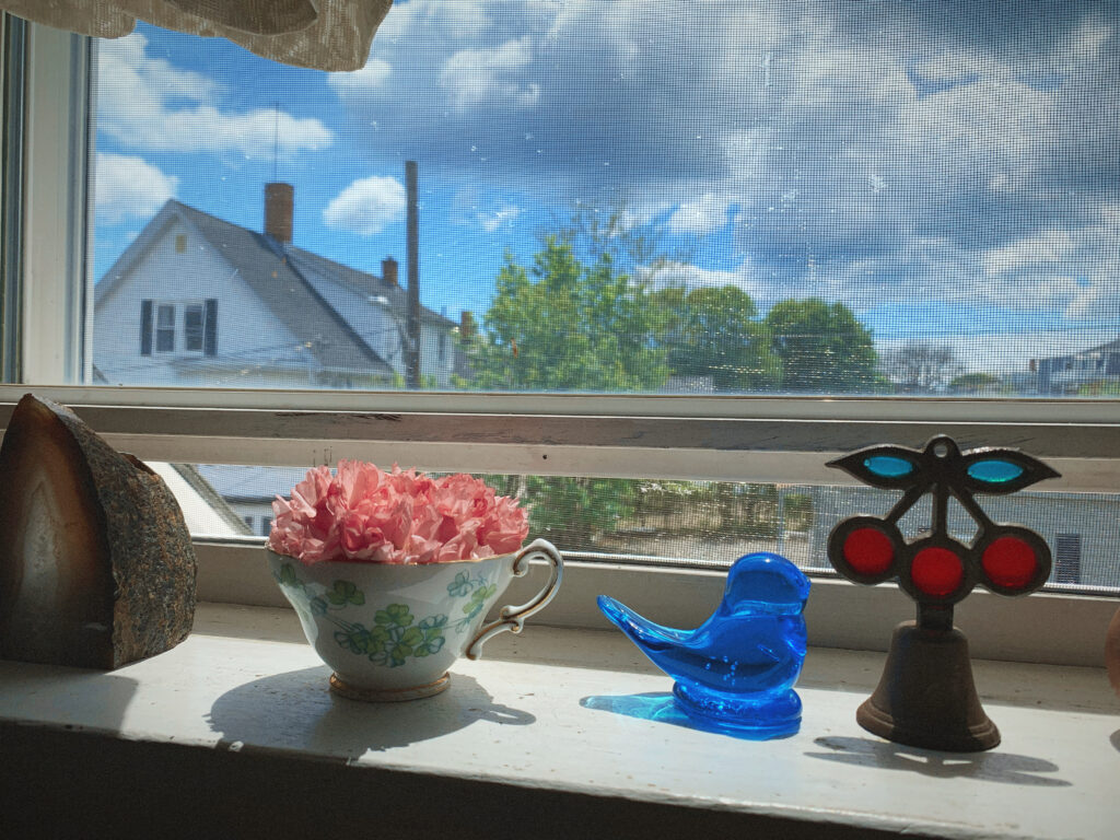 A sunny windowsill with four objects on it. From left to right: a chunky geode rock, a shamrock-pattern teacup holding a spray of cherry blossoms, a glass-blown bluebird of happiness, and a small bell with a stained glass handle in the shape of three cherries. The sky in the background is blue with big puffy white clouds.