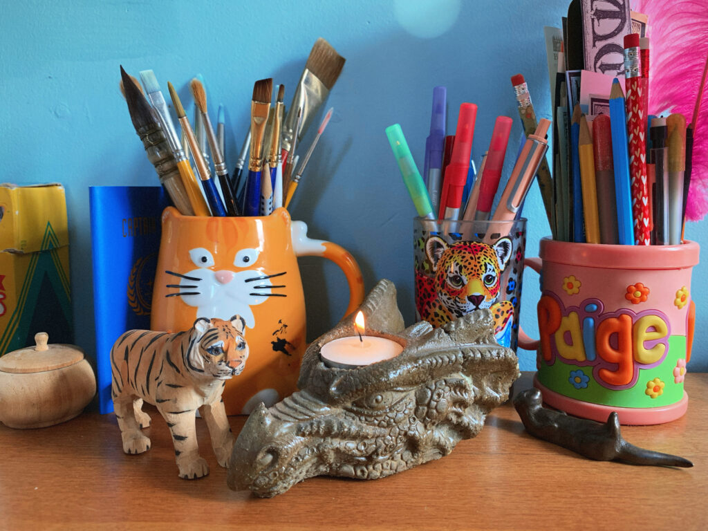 A small painted wooden tiger and a stone candle holder shaped like a dragon's head and holding a lit tealight. Behind the figurines are three cups filled with paintbrushes, pens, and pencils. The left cup is shaped like an orange cat, the middle cup has a Lisa Frank rainbow leopard on it, and the third cup says PAIGE in multicolored letters.