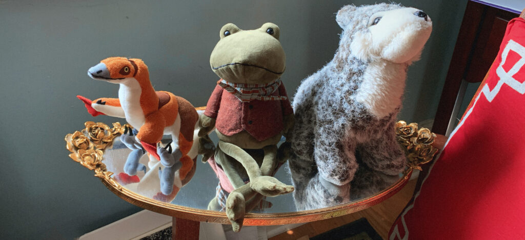 Three stuffed animals sitting on an oval mirror. From left to right: An orange dinosaur-bird, the Jellycat Riverside Rambler frog wearing an oxblood vest and striped scarf, and a grey wolf resting on her haunches. The mirror is rimmed in gold and has one handle on either side. Each handle is fashioned like a bunch of roses.