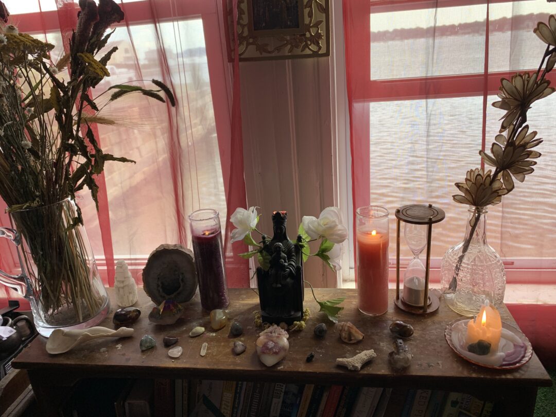 Photo of a shrine to the Black Madonna on top of a bookshelf. The ocean is visible through the sheer red curtains on the two windows behind the shrine. Several red candles are burning among offerings of shells and curios. There are vases of dried flowers on either end of the altar.